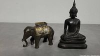 Photo showing the full view of the cast iron and metal elephant coin bank and seated Buddha statue side by side on a neutral surface.
