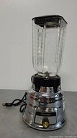 Front view of Osterizer blender showing chrome base with control knob and attached glass jar with black lid.