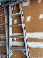 Photo showing a 6-foot aluminum ladder leaning against a wall in a garage or workshop with visible screws and patchwork on the wall.