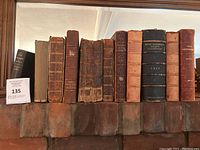 Full view of 12 leather-bound antique books lined up on a shelf. Visible wear and patina on all covers.