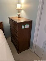 Side view of antique tall boy chest of drawers showing height and finish beside a bed and lamp on top.