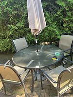 Full view of round glass-top outdoor patio table with central umbrella pole and four metal framed chairs with mesh backing and seats around it, placed on a paved backyard patio with greenery in background.