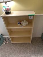 Front view of beige solid wood bookshelf with three shelves, with a lamp and decorative items nearby but not included.