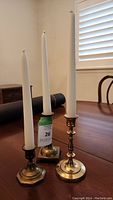 Three brass candle holders on a wooden table, each holding a white taper candle. Different shapes and patinas visible.