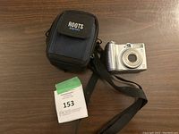 Silver Roots digital camera with black strap along side black and navy blue padded case marked Roots Digital placed on wooden surface.