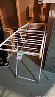 White folding laundry drying rack with multiple horizontal rods standing upright in a storage space.