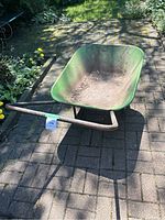 Photo showing a large green metal wheelbarrow with wooden handles and some dirt inside the tray, placed on an outdoor brick patio with garden background.