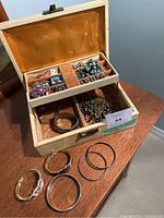 Photo showing the wooden jewelry box opened on a table displaying colorful brooches, rings, beaded jewelry inside, and seven assorted bangle bracelets placed around the box.