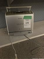 Side angled view of silver Canon Selphy photo printer with front paper output slot and power cord, showing model name and branding.
