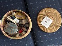Round multi-colored woven basket with lid removed showing assorted sewing items inside including scissors, thread spools, pins, fabric, and other sewing accessories.