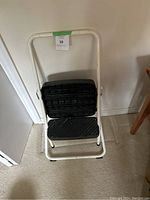 White metal frame step stool with two black textured steps and handle bar shown against a wall and near furniture on carpet.
