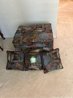 Front view of the tapestry ottoman and three matching pillows arranged on the floor showing detailed garden scene upholstery.