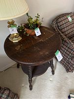 Full view of vintage wooden side table with round top, lamp, and plant on top, showing condition and setting.