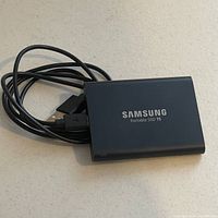 Samsung Portable SSD T5 with USB 3.0 cable on white surface showing Samsung logo and T5 branding on front side of the drive.