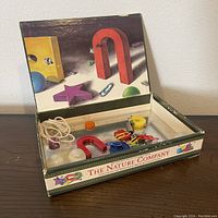 Open box showing the inside with various magnets including a large red horseshoe, paper clips, balls, yellow circular magnet, and a string on a magnetic base inside a printed box.
