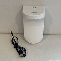 Front view of white Fohm touchless soap dispenser with USB charging cable coiled beside it.