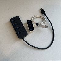 Two USB hubs on a white surface: one small Sabrent 4-port hub with silver frame and one larger black 4-port hub with individual port covers. Both have attached USB cables.