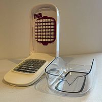 OXO vegetable chopper showing white hinged lid with white knob pusher and stainless steel blade grid, plastic basin next to unit showing a small chip at edge