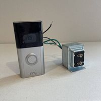 View of Ring Video Doorbell 3 front side showing camera and button, alongside a metallic hardwired transformer with attached wiring.