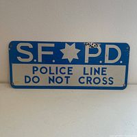 Photo showing front of blue and white metal SFPD Police Line Do Not Cross sign with star emblem and large text.