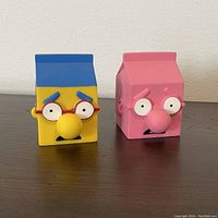 Front view of two MILKHOUSE soft vinyl sculptures with cartoon faces inspired by The Simpsons, showing detailed facial features and colors.