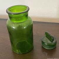 Small green glass bottle and separate matching stopper shown on wooden surface.