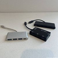 Three USB-C hubs displayed on a white surface including a silver hub with three USB-A ports and one USB-C passthrough port, a black hub with three USB-A ports and an Ethernet port with Dirago branding, and another black hub with three USB-A ports and a USB-C passthrough port.