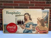 Full view of vintage 1948 Coca Cola litho cardboard sign with two pinup girls and Coca Cola bottles on tray, with word Hospitality and Coca-Cola round logo.