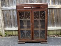 Front view of the antique wooden waterfall cabinet showing two sliding drawers on top and two glass doors with metal design overlays closed.