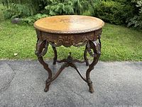 Full view of the antique round hand-carved table with marquetry top on an outdoor paved surface.