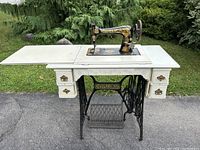 Full frontal image of the entire antique Singer sewing machine attached to a wooden cabinet. the cabinet is painted white with gold/blue floral accents and has iron legs with foot pedal base.