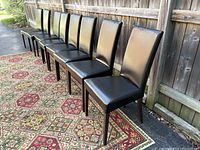 Eight black padded dining chairs without covers lined up outside on a patterned carpet, side view.