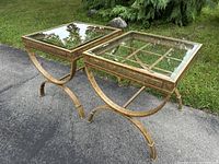 Two metal framed square accent tables with glass tops shown outdoors on driveway with greenery in background.