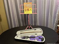 Photo showing the pink foldable music stand with the yellow instructional violin book on it and the gray padded violin case open with the purple violin and bow inside.