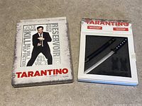 Photo showing box set of Quentin Tarantino DVD Ultimate Collection with cover featuring film titles and a man holding a gun, placed next to packaging with mini samurai sword and stands.