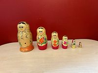 Six vintage Russian Matryoshka nesting dolls arranged from largest to smallest on a table against a red background. Dolls are wooden and hand-painted with various floral and folk designs.