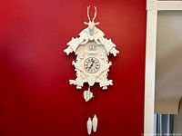 Full view of the white carved cuckoo clock mounted on red wall showing the overall size and design including deer head, birds and leaves.