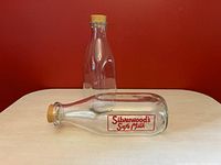 Two clear glass pint milk bottles with red applied colour label reading 'Silverwood's Safe Milk', standing and lying on a beige surface against a red background, both sealed with original rubber stoppers.