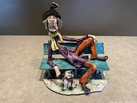 Front view of the ceramic statue showing the man reclining on the teal bench with dog and bird on his hat.