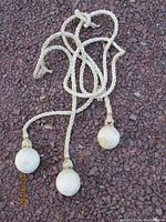 Full view of the handmade gaucho bola showing three braided cords with three weighted balls attached at the ends, laid on gravel surface.