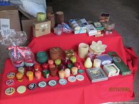 Photo of large assortment of candles spread out on a red surface showing various sizes, colors, and types of candles, with some boxed items in the background.