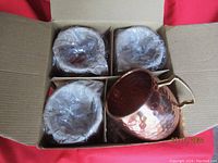 Four new hammered copper Moscow Mule mugs in original packaging with plastic liners, set inside an open cardboard box.