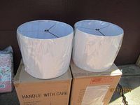 Two new white drum lamp shades shown sitting on cardboard boxes, wrapped in plastic, with metal support frames visible inside.