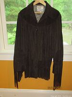 Front view of brown suede Pioneer Wear western jacket hanging on hanger near window, showing long fringe detailing from chest to bottom hem and sleeves.