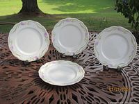 Four white porcelain dinner plates with scalloped edges and purple floral decoration on the rim, displayed standing on a metal table outdoors.