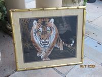 Framed silk tiger art piece depicting a tiger stepping forward with an aggressive facial expression, signed in lower corner, bamboo-style gold frame.