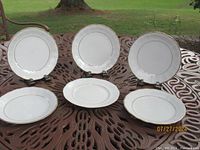 Photo showing six white porcelain plates with gold or decorative rims displayed on a metal outdoor table. Five plates are from Walbrzych, and one is from Korona, both Polish manufacturers.