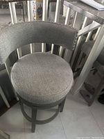 Full view of one gray upholstered bar stool showing round cushioned seat, curved backrest with silver nailhead trim and wooden legs with footrest ring.