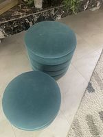 Two teal blue round velvet-like fabric upholstered storage ottomans with removable lids, stacked on tiled floor near a marble surface and a carpet.