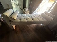 Gray velvet chaise lounge couch placed indoors on brown wooden flooring, showing tufted seat and wooden legs along with bolster pillow and headrest pillow.
